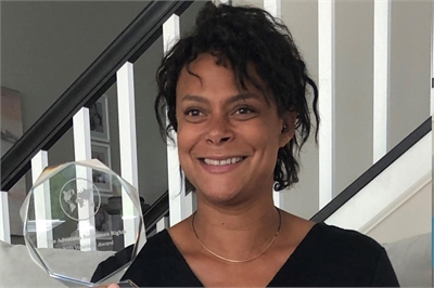 [ai] A woman with curly hair smiling while holding a crystal award in a well-lit room with white walls and a staircase in the background.